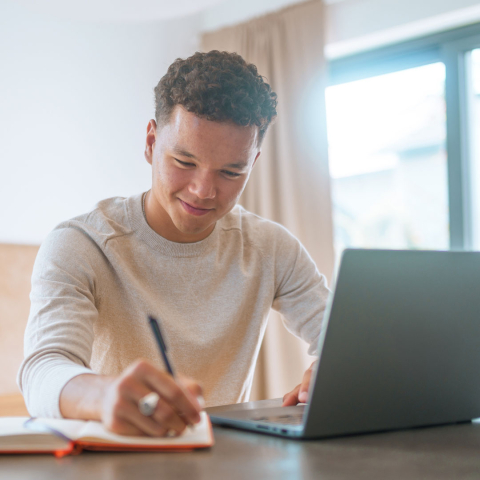 Teen boy writing in a journal while laptop is open