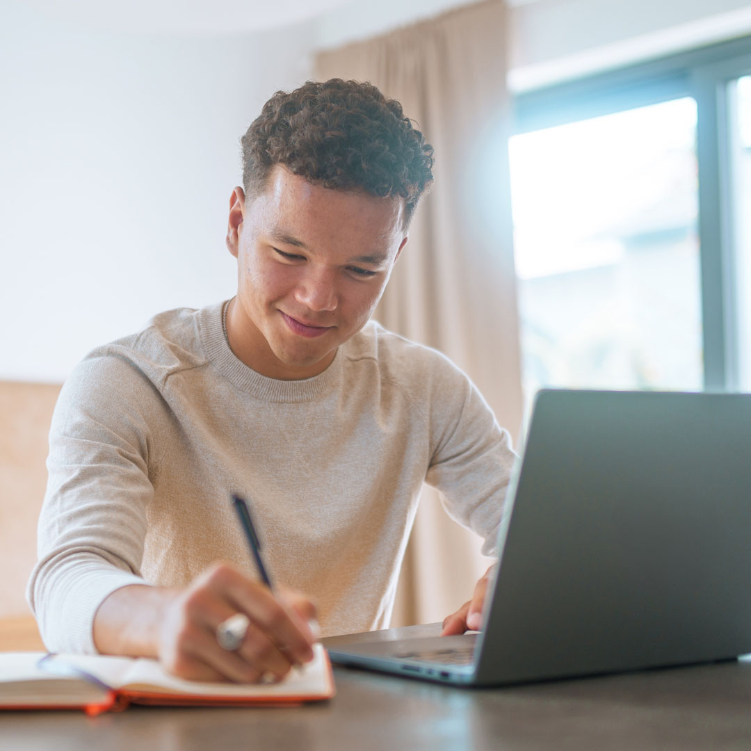 Teen boy writing in a journal while laptop is open